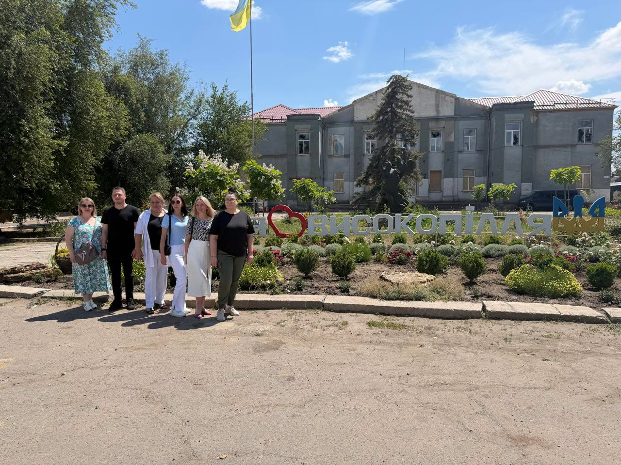 Група людей позує для фото на тлі великого патріотичного напису з назвою громади, прикрашеного декоративним серцем та синьо-жовтими літерами «UA». Позаду доглянутої клумби з квітами та зеленими кущами розташована двоповерхова будівля, над якою на високому флагштоці майорить державний прапор України.