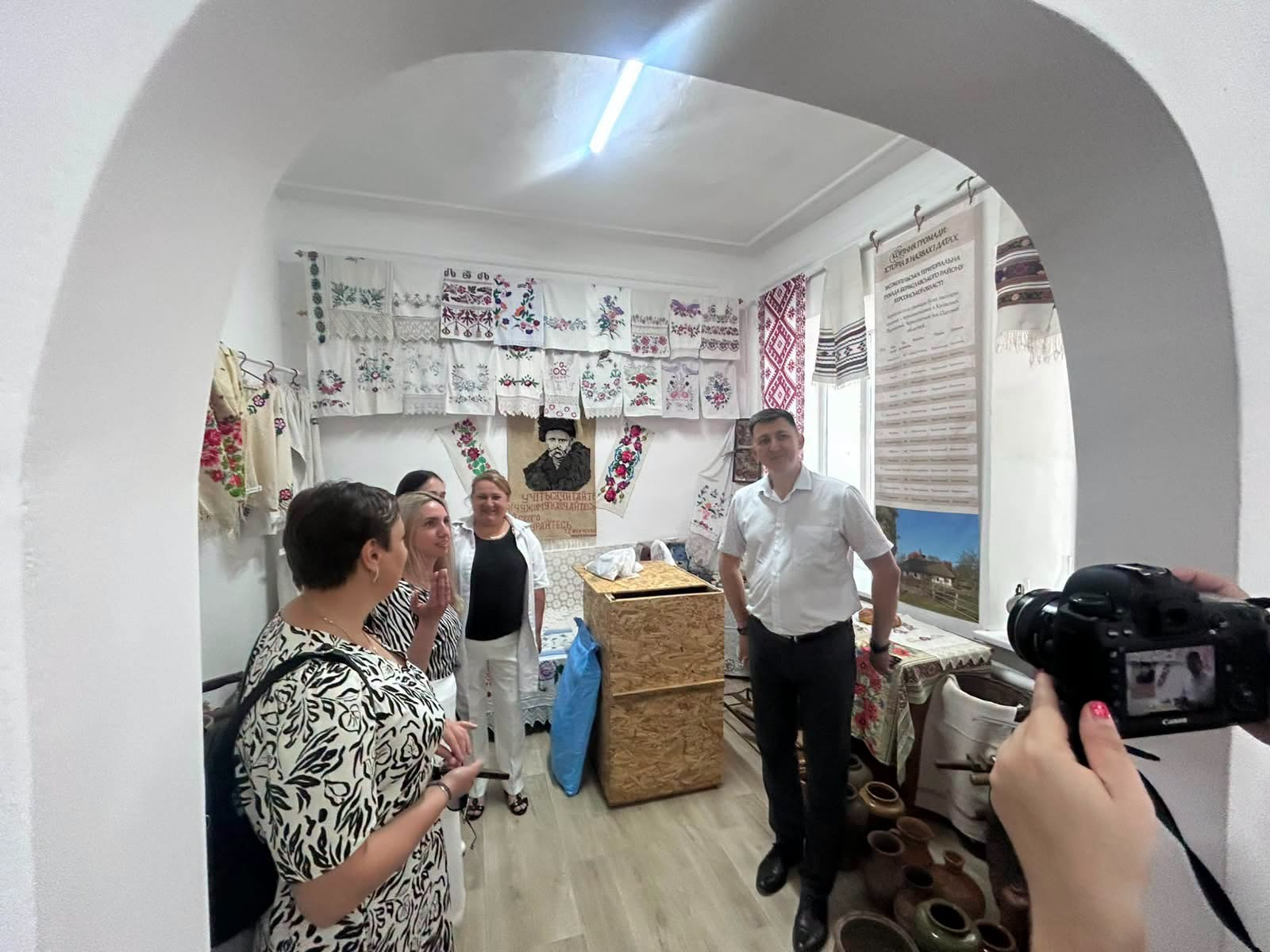 Група людей перебуває в музейній кімнаті з традиційним українським інтер'єром, оглядаючи експонати. На стінах розвішані вишиті рушники та сорочки, по центру розміщено портрет Тараса Шевченка, а на передньому плані справа людина знімає те, що відбувається, на цифровий фотоапарат.