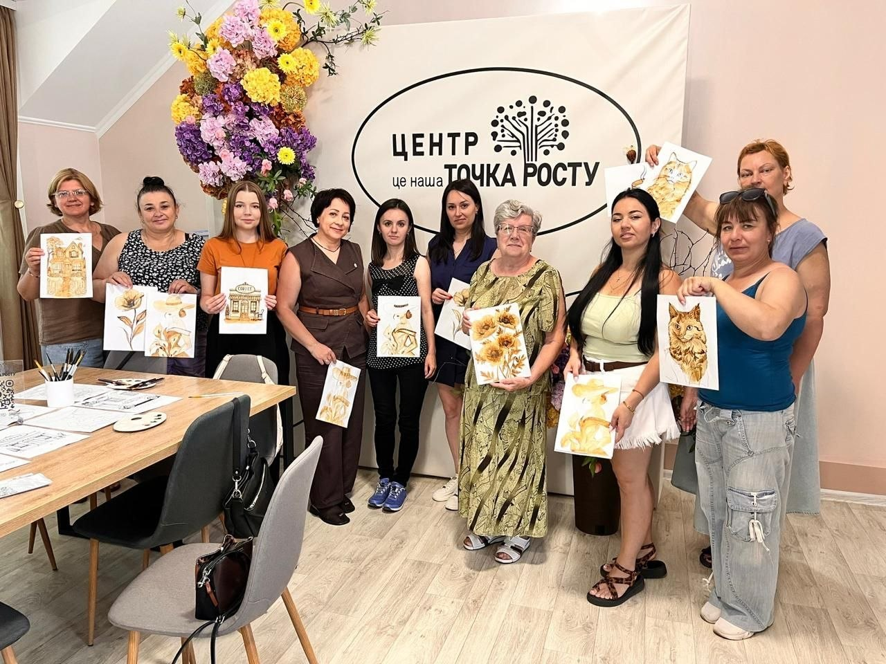 Група жінок позує для спільного фото в світлій кімнаті, тримаючи в руках свої готові малюнки в однакових золотаво-коричневих тонах із зображеннями квітів, котів та будівель. Вони стоять біля великого банера з написом «ЦЕНТР це наша ТОЧКА РОСТУ», прикрашеного яскравою квітковою інсталяцією, а ліворуч видно частину робочого столу зі стільцями.