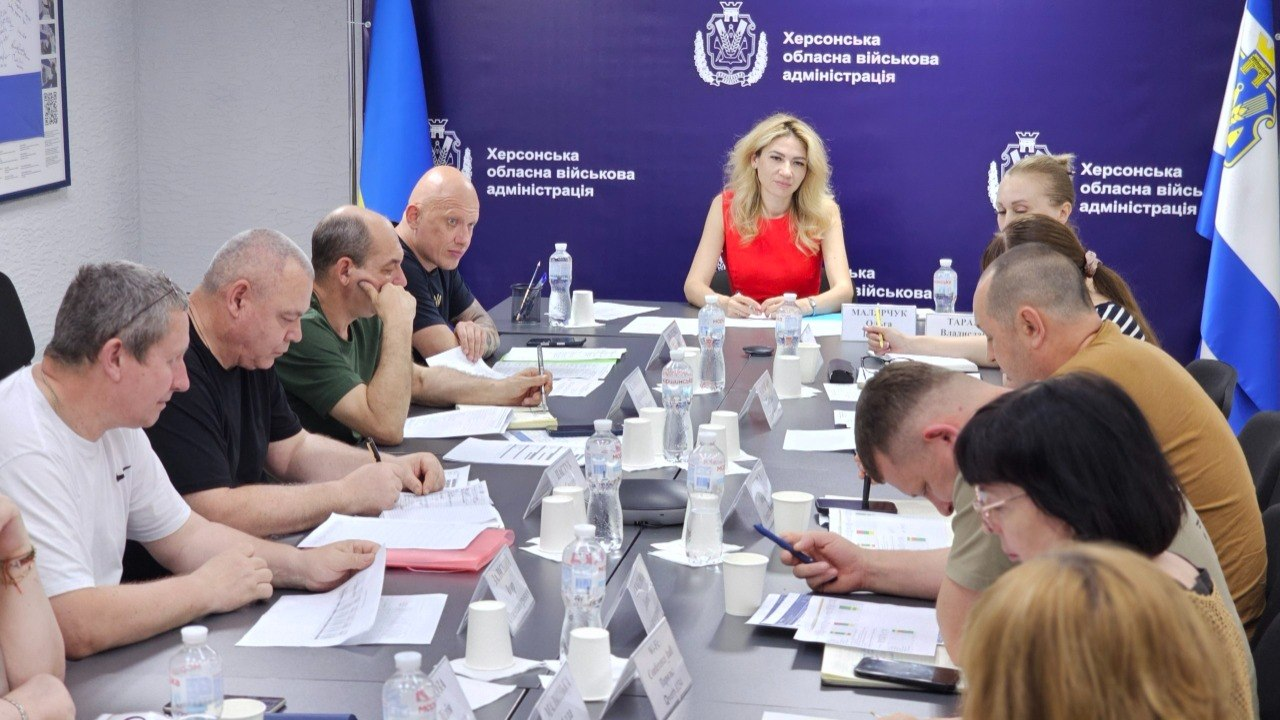Група людей сидить навколо великого столу під час робочої наради в офіційному кабінеті. На синьому фоні позаду них розміщено герб, а ліворуч і праворуч стоять державні прапори України. На столі перед учасниками зустрічі лежать документи, блокноти, ручки та пляшки з водою.