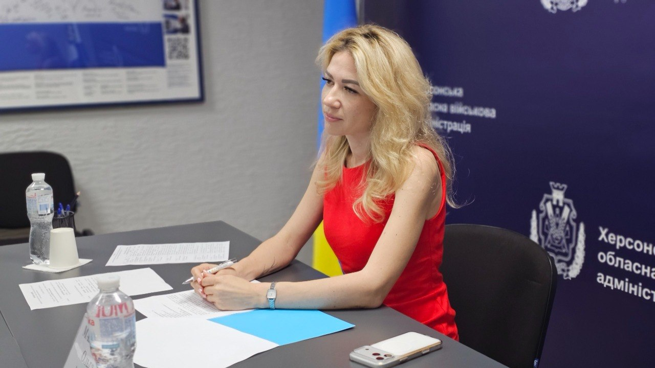 Жінка у яскравій червоній сукні сидить за робочим столом в офіційному кабінеті на тлі синьої стіни з гербом. Вона тримає в руках ручку, а перед нею на столі лежать паперові документи, папка, мобільний телефон та пляшка води.