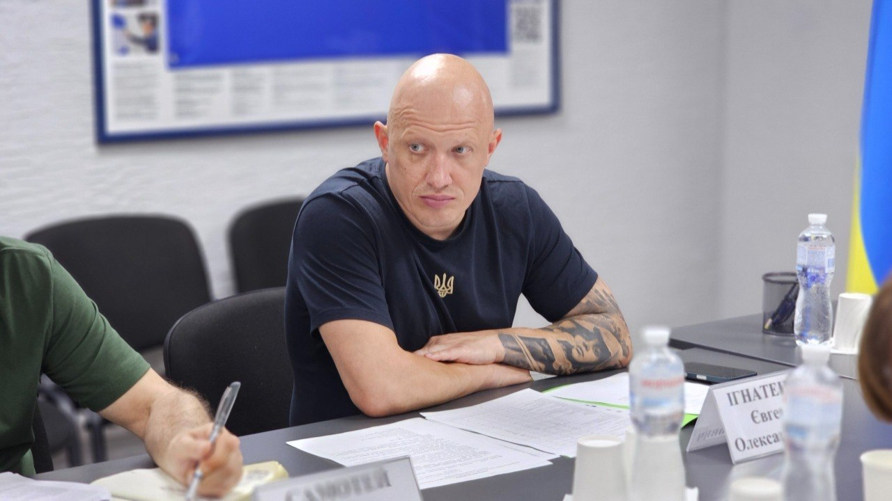 Чоловік у темно-синій футболці з малим золотистим гербом України сидить за робочим столом під час наради. Він склав руки перед собою, а перед ним лежать документи, таблички з іменами та пляшки з водою.