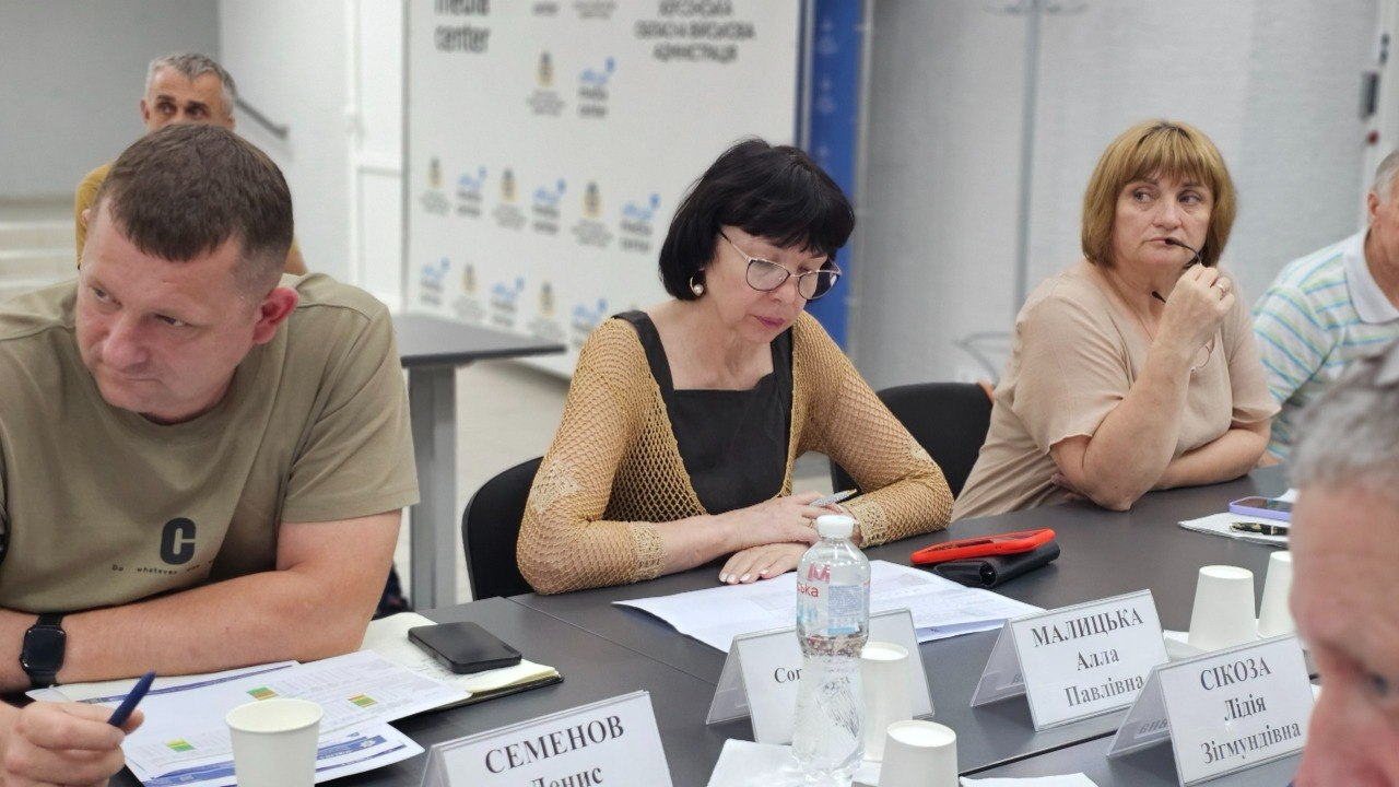 Учасники робочої зустрічі сидять за великим столом у кабінеті та вивчають документи. Перед ними розміщені паперові матеріали, настільні таблички з іменами, пляшка води та пластикові стаканчики, а на задньому плані видно світлу стіну з офісними логотипами.
