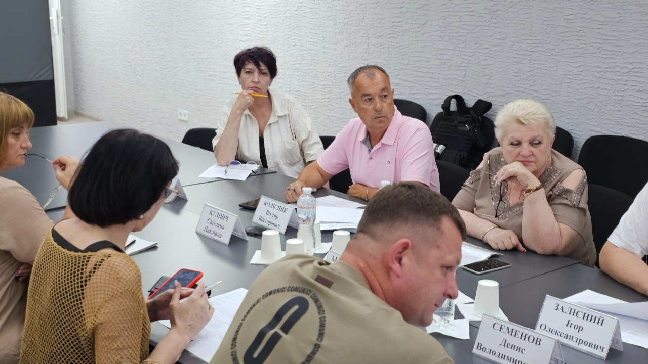 Учасники робочої зустрічі сидять за великим столом в офіційному приміщенні та слухають доповідача. Перед ними лежать паперові документи, мобільні телефони, а також розставлені настільні таблички з іменами та пластикові стаканчики.