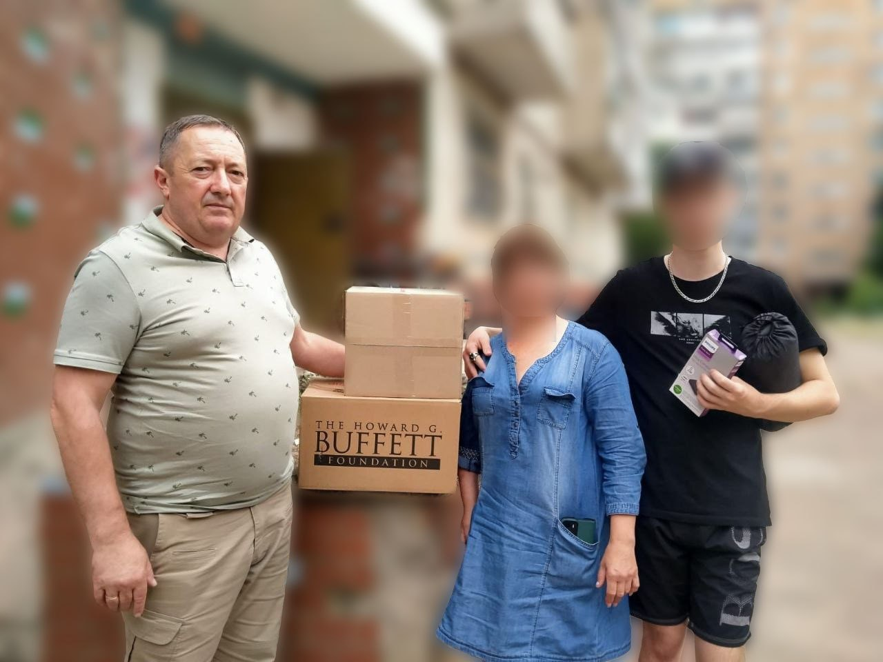 Три людини стоять на вулиці біля житлового будинку під час отримання допомоги. Чоловік та жінка тримають дві картонні коробки, на одній з яких розміщено напис фонду, а хлопець поруч тримає в руках невеликі пакунки. Обличчя жінки та хлопця на зображенні розмиті.