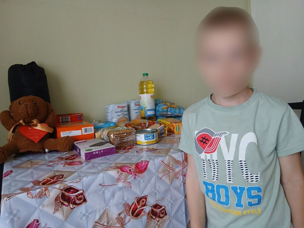 Хлопчик у світлій футболці з написом стоїть у кімнаті поруч зі столом, на якому розкладено гуманітарну допомогу. На вкритому скатертиною столі лежать різноманітні продукти харчування: пляшка олії, крупи, консерви, печиво, а також коробки та плюшеве ведмежа. Обличчя дитини на фото розмите.