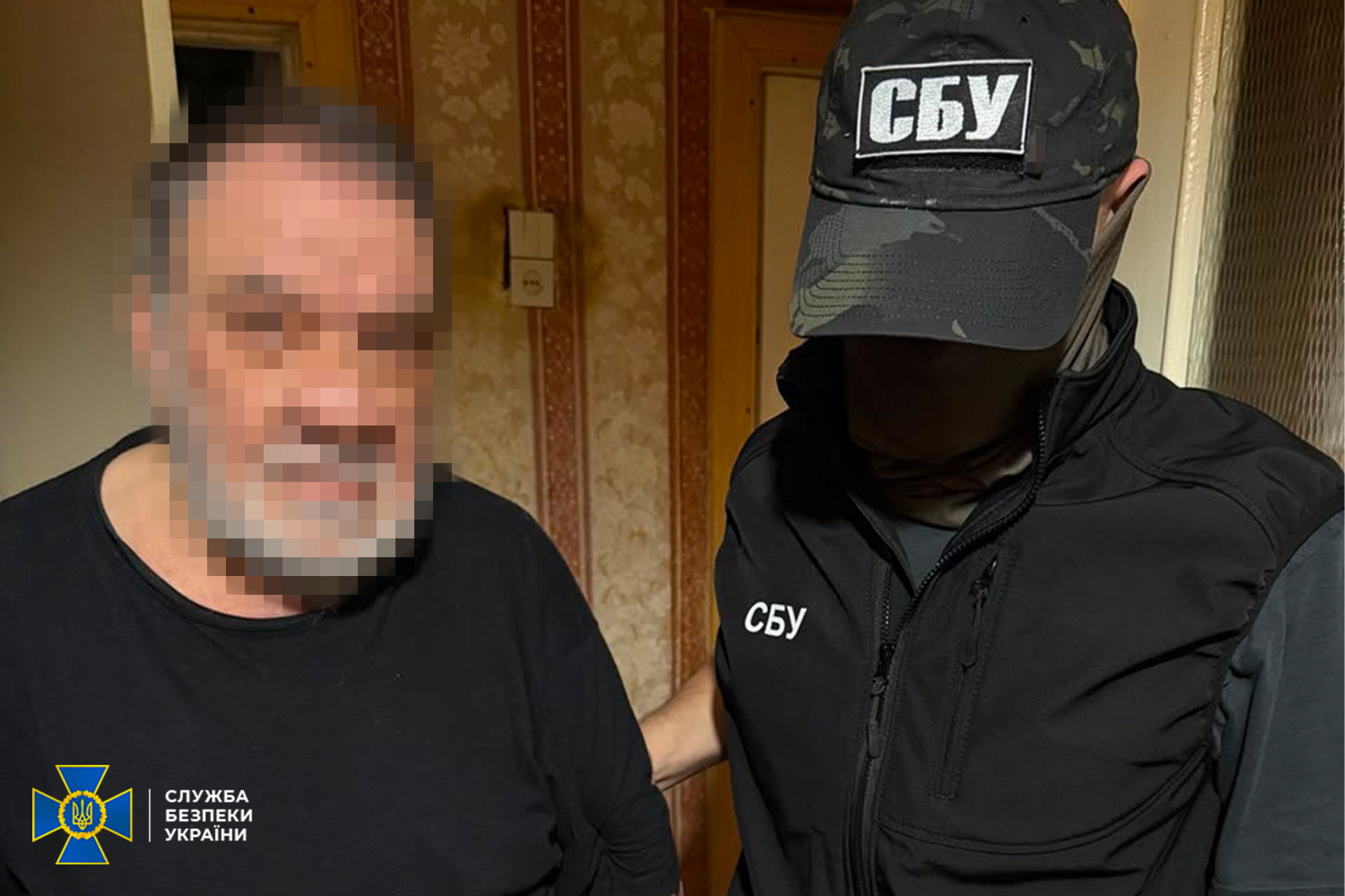 Чоловік у чорному одязі із розмитим обличчям стоїть у приміщенні поруч із правоохоронцем. Співробітник у камуфляжній кепці та жилеті з написами «СБУ» тримає чоловіка за руку, при цьому обличчя представника служби повністю приховане кепкою. У лівому нижньому кутку фото розміщено офіційний логотип Служби безпеки України.