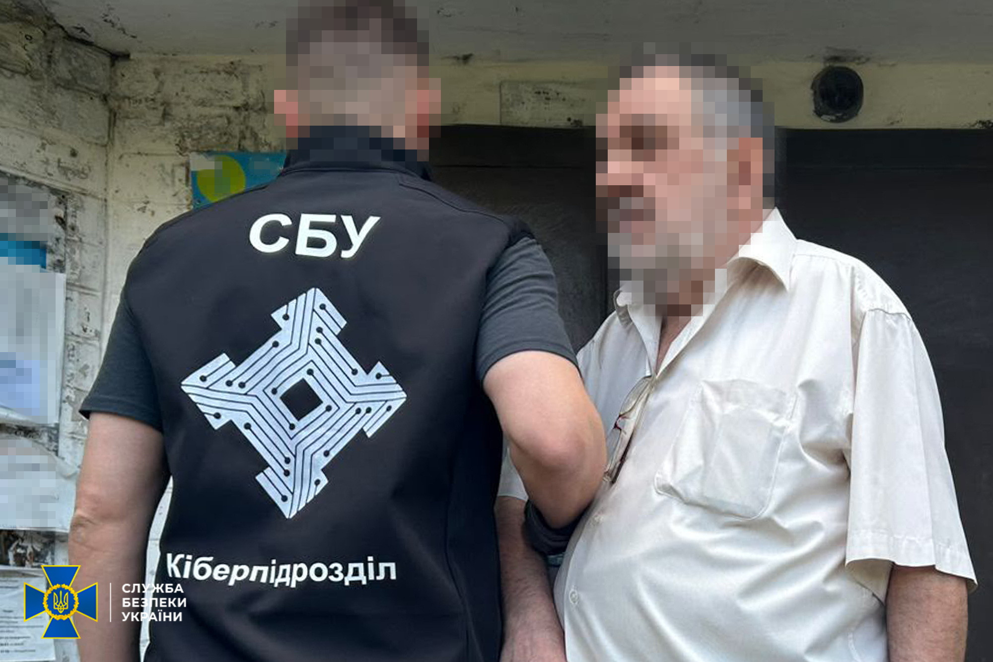 Чоловік у світлій сорочці з розмитим обличчям стоїть на вулиці поруч із правоохоронцем, який повернутий спиною до камери. Співробітник одягнений у жилет із написом «СБУ», емблемою та написом «Кіберпідрозділ», а його обличчя також розмите. У лівому нижньому кутку фото розміщено офіційний логотип Служби безпеки України.
