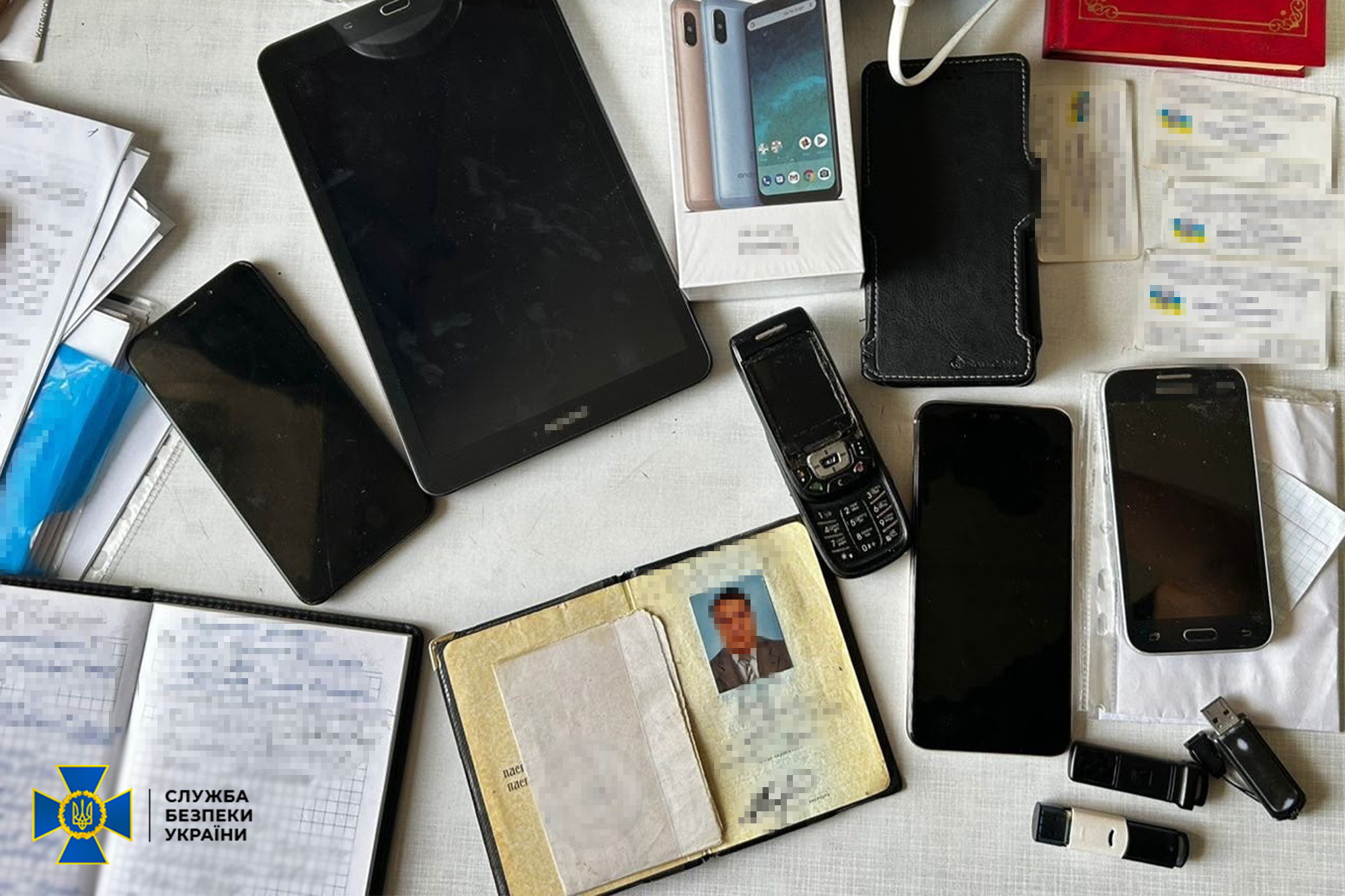 На світлому столі розкладені речові докази: кілька мобільних телефонів, кнопочний телефон, планшет, коробка від смартфона, USB-флешки та паперові документи. Серед документів видно розгорнуте посвідчення з фотокарткою чоловіка та кілька пластикових карток із розмитим текстом, а у лівому кутку розміщено логотип Служби безпеки України.