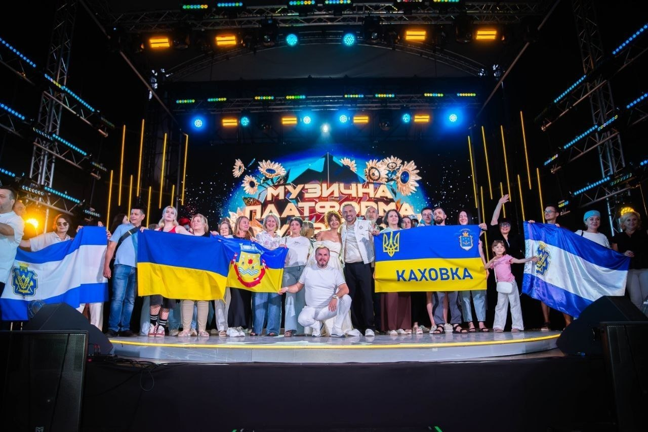 Велика група людей стоїть на концертній сцені з яскравим підсвічуванням на тлі екрана з написом «МУЗИЧНА ПЛАТФОРМА». Учасники тримають розгорнуті прапори України, Херсонської області, Каховки та Каланчака, позуючи для спільного фото.