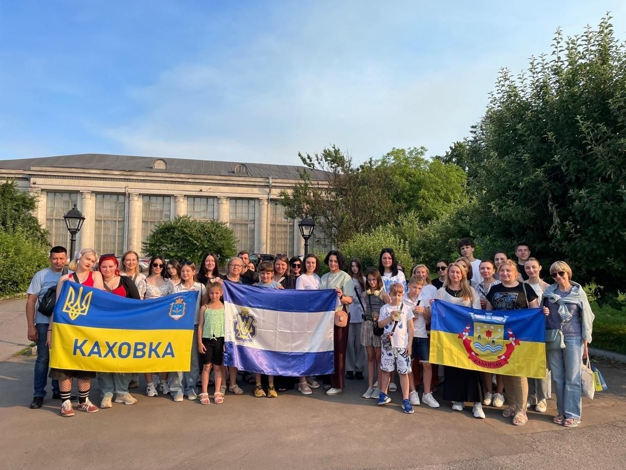 Група дорослих та дітей позує для спільного фото на вулиці на тлі великої будівлі з колонами та зелених дерев. У руках вони тримають три розгорнуті прапори: ліворуч — синьо-жовтий прапор із написом «КАХОВКА», у центрі — прапор Херсонської області, а праворуч — синьо-жовтий прапор із гербом Каланчака.