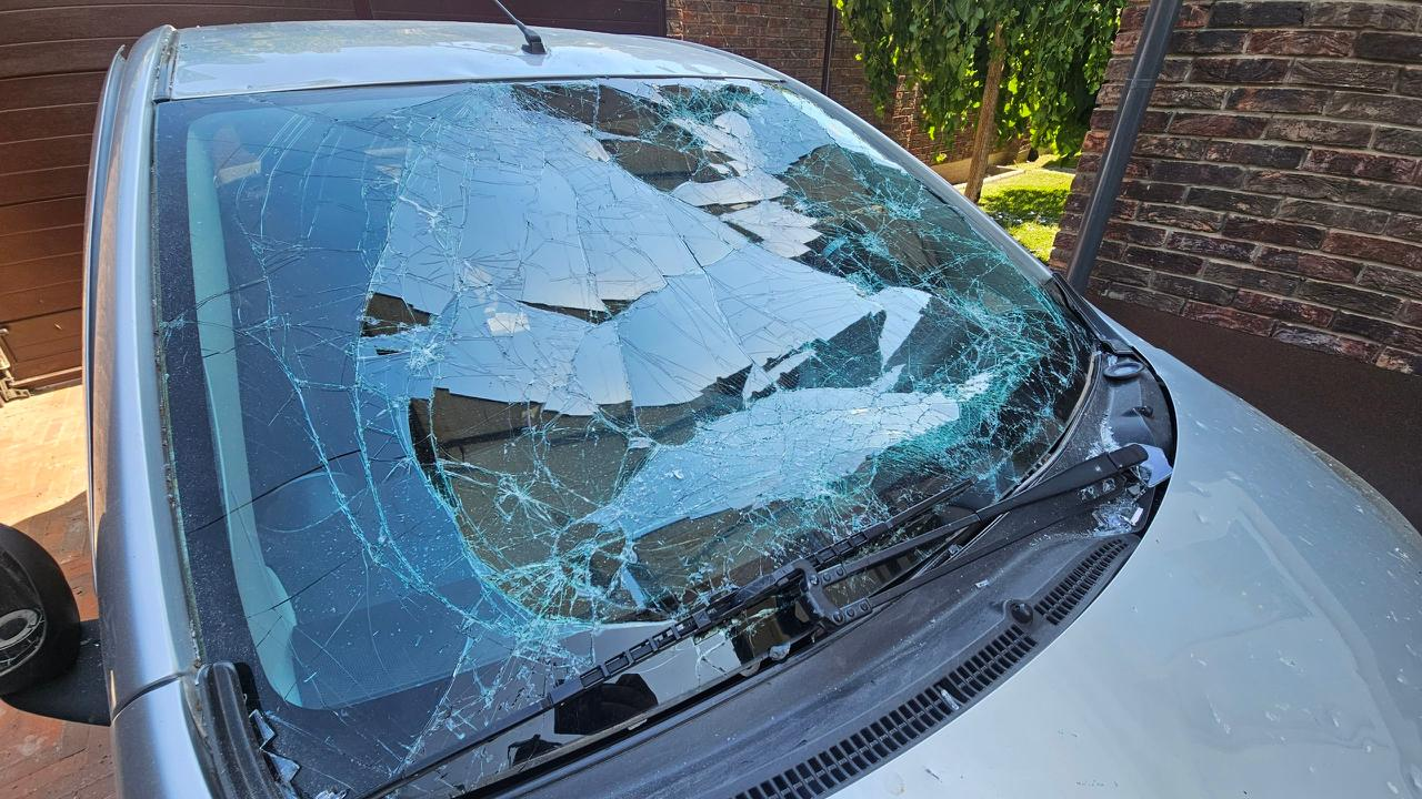 Сріблястий автомобіль із повністю розтрощеним лобовим склом, яке вкрите густою сіткою глибоких тріщин та частково прогнулося всередину салону. Машина стоїть на вулиці поруч із цегляною стіною будівлі та коричневими гаражними воротами.