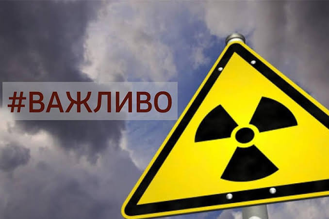 Трикутний жовтий знак радіаційної небезпеки із чорним символом трилисника, закріплений на стовпі. Праворуч від нього на тлі темного грозового неба розташований напис «#ВАЖЛИВО» бордового кольору на напівпрозорій плашці.