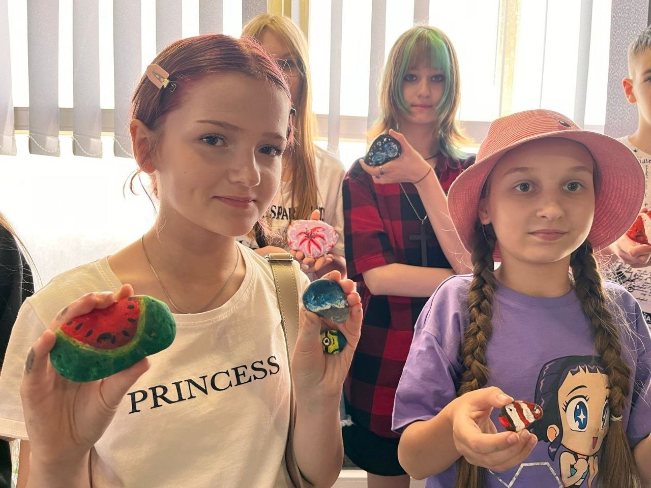 Група дітей та підлітків стоїть у приміщенні біля вікна з жалюзі та демонструє на камеру розписане вручну каміння. На передньому плані дівчина у білій футболці тримає камінь із малюнком кавуна, а інші учасники показують камінці з морськими пейзажами, квітами та абстрактними візерунками.