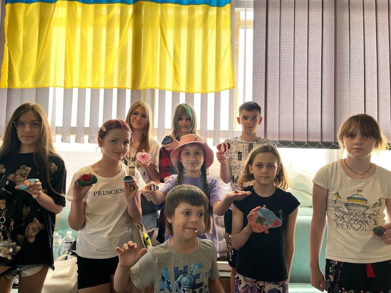 Група дітей та підлітків позує для спільного фото у приміщенні на тлі вікна з жалюзі та державного прапора України. Вони тримають у руках та демонструють на камеру невеликі розписані вручну камені із різноманітними яскравими малюнками.