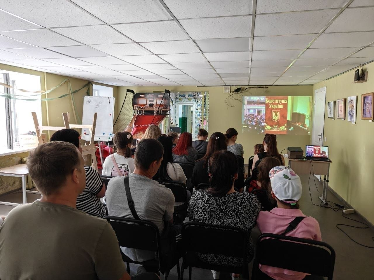 Група людей сидить на стільцях у довгому приміщенні та дивиться на стіну, куди через проєктор транслюється зображення з написом «Конституція України» та державним гербом. Ліворуч біля вікна видно фліпчарт, а на правій стіні розвішані невеликі картини в рамках.
