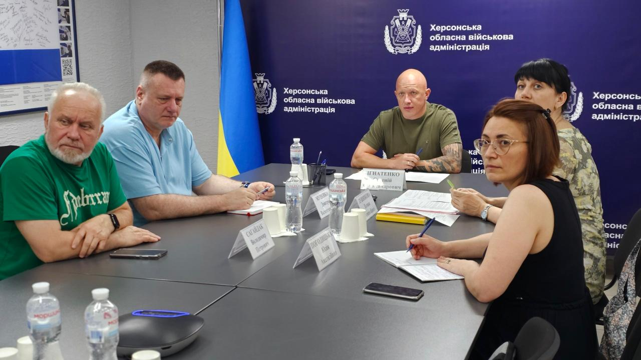 Учасники зустрічі сидять за великим столом під час офіційного заходу в кабінеті. Позаду них розміщено синій банер із гербом, ліворуч стоїть державний прапор України, а на столі перед присутніми розставлені таблички з іменами, паперові документи, блокноти та пляшки з водою.