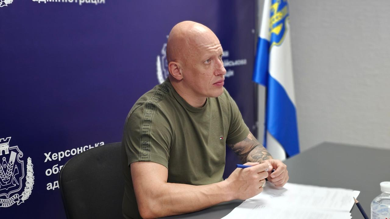 Чоловік міцної статури з поголеною головою та татуюванням на лівій руці сидить за столом на тлі синього банера з гербом Херсона та прапора України, тримаючи в руках ручку.