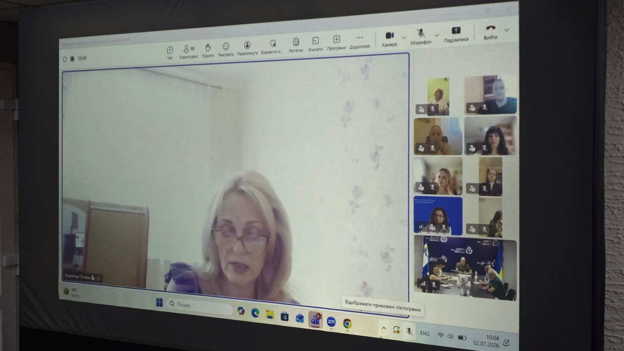 Великий проєкційний екран відображає інтерфейс онлайн-наради, де у головному вікні виступає жінка в окулярах, а праворуч розташована панель із меншими вікнами інших учасників відеозустрічі.