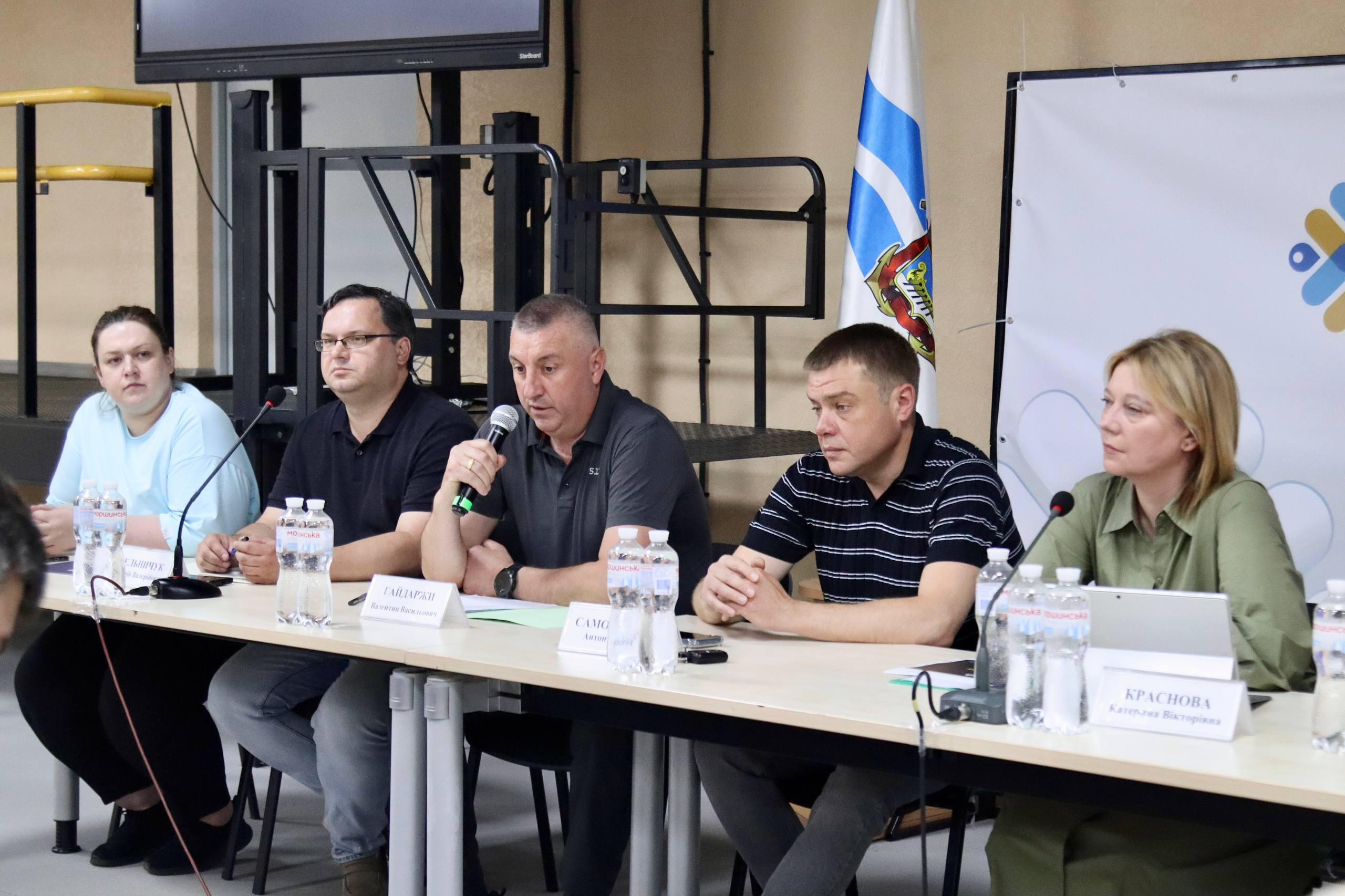 П'ятеро людей сидять за довгим столом під час офіційного заходу, один із чоловіків говорить у мікрофон, на столі стоять таблички з іменами та пляшки з водою, а на задньому плані видно прапор і технічне обладнання.