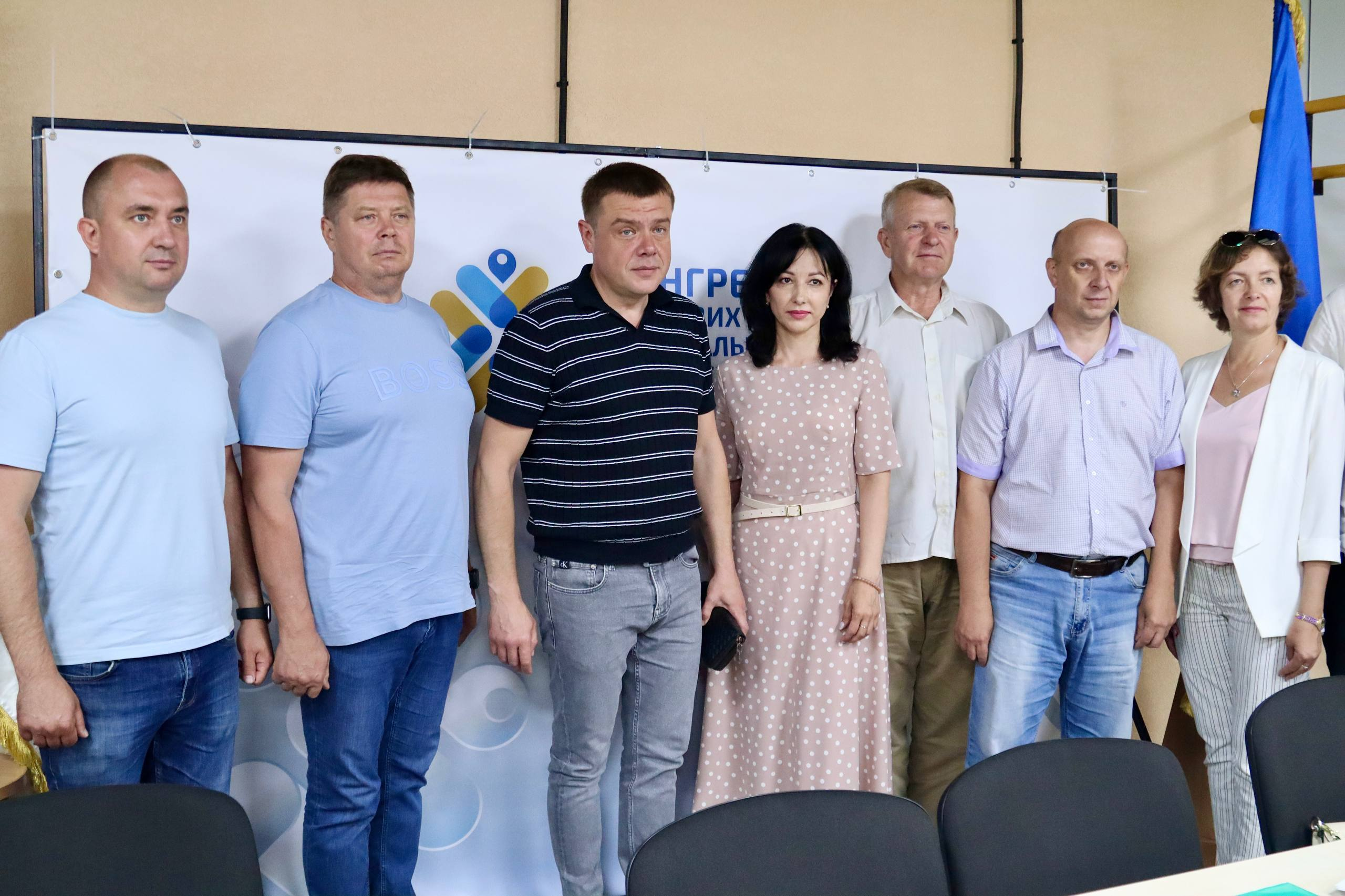 Група людей у літньому повсякденному та офіційно-діловому одязі стоїть в один ряд для спільного фото на тлі банера з логотипом та синьо-жовтого прапора.