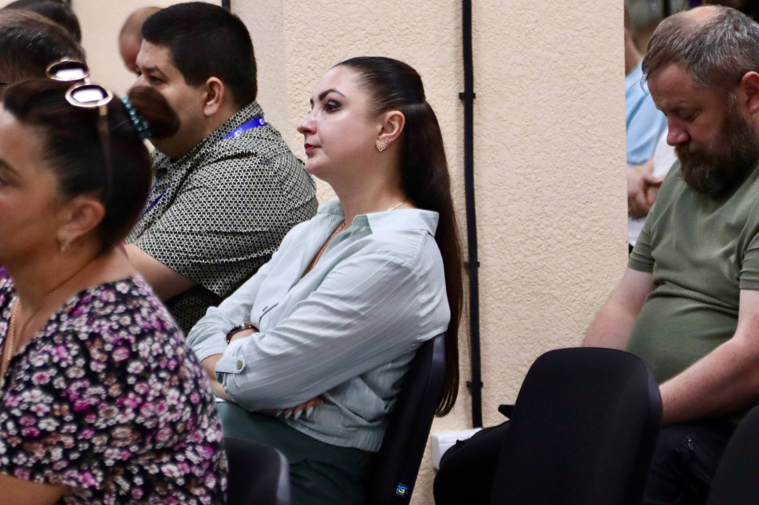 Жінка зі зібраним у хвіст довгим темним волоссям сидить у залі серед інших слухачів, склавши руки на грудях та уважно дивлячись у бік президії.