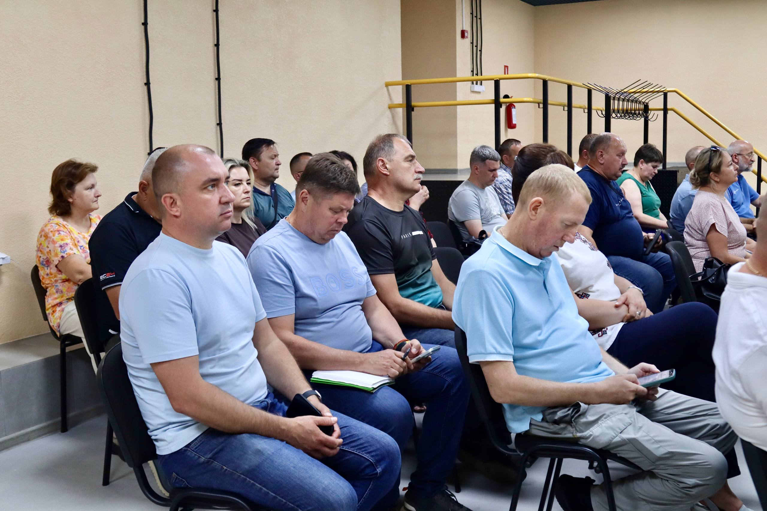 Група людей сидить на стільцях у залі під час заходу, слухаючи виступ та занотовуючи інформацію у свої телефони та блокноти.