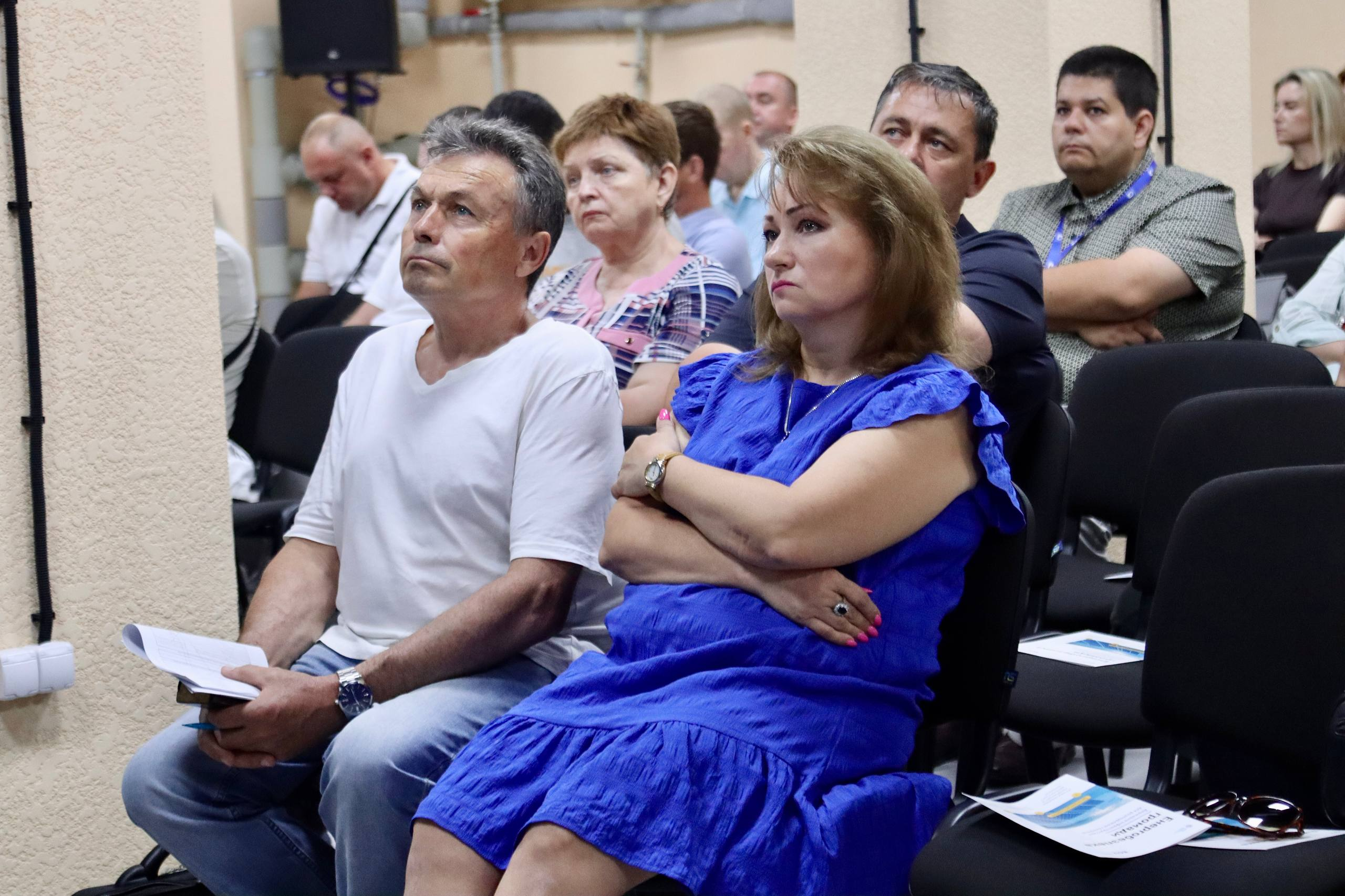 Жінка у синій сукні та чоловік у білій футболці сидять у залі серед інших учасників заходу та зосереджено дивляться вперед.