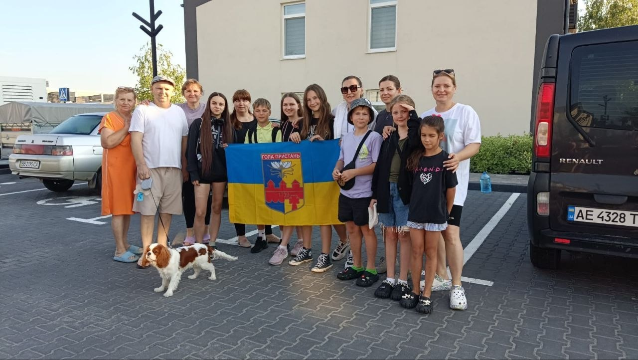Група дорослих та дітей стоїть на автомобільній парковці, тримаючи прапор міста Гола Пристань, поруч із ними на повідку стоїть невеликий біло-рудий собака.
