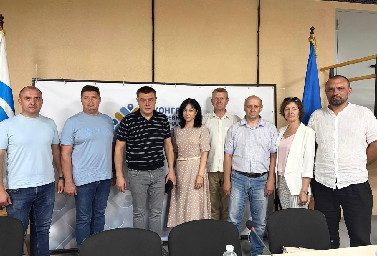 Група з восьми людей у повсякденному та діловому одязі стоїть у шерензі в офіційному приміщенні на тлі білого банера. По обидва боки від них встановлено прапори, а на передньому плані розташовані офісні стільці та стіл.