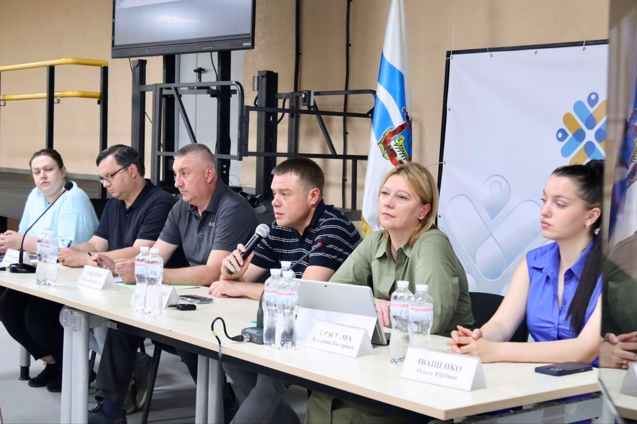 Група людей сидить за довгим білим столом під час офіційного заходу. Чоловік у центрі говорить у мікрофон, перед учасниками стоять пляшки з водою та таблички з іменами, а позаду видно прапор і білий банер із логотипом.