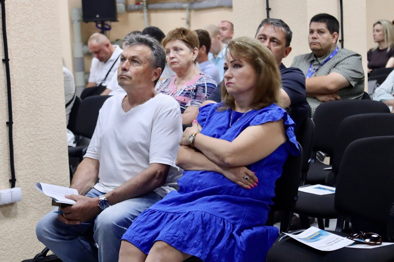 Група людей сидить на чорних стільцях у конференційній залі під час офіційного заходу чи презентації. На передньому плані чоловік у білій футболці та жінка в синій сукні уважно дивляться вперед, а на сусідніх сидіннях розкладені інформаційні друковані матеріали.