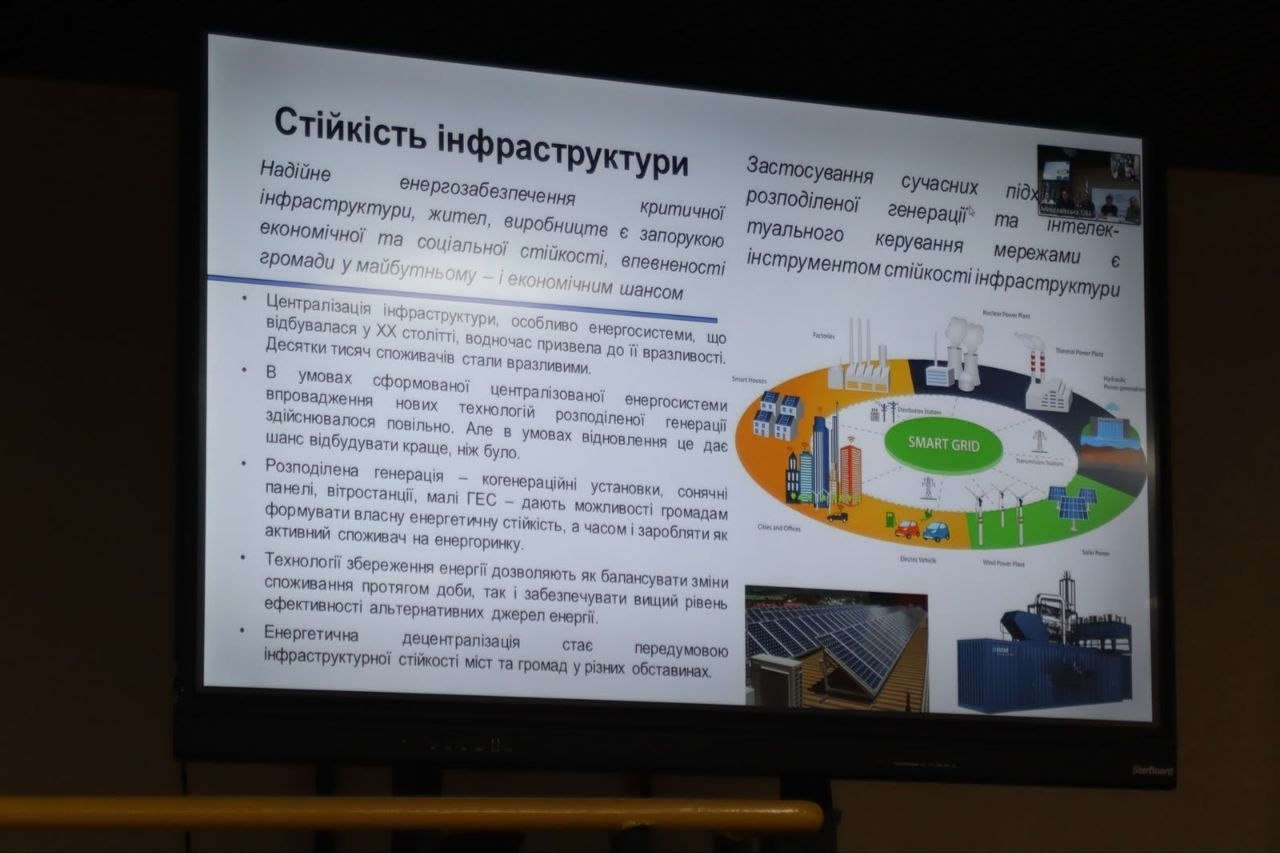 Презентаційний слайд на великому екрані з заголовком «Стійкість інфраструктури». Слайд містить блоки українського тексту про енергозабезпечення та децентралізацію, маркований список, ілюстрацію схеми розумної електромережі («SMART GRID») з вітряками та будинками, а також два невеликі фотоелементи з сонячними панелями та промисловим обладнанням.