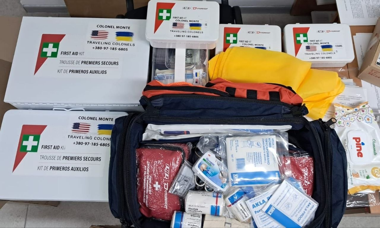 Набір гуманітарної медичної допомоги, що складається з великої відкритої сумки-аптечки, заповненої перев'язувальними матеріалами та медикаментами, а також кількох закритих білих пластикових боксів різного розміру. На коробках розміщено наліпки з медичним хрестом, прапорами США та України, а також написами «FIRST AID KIT» та «COLONEL MONTE».