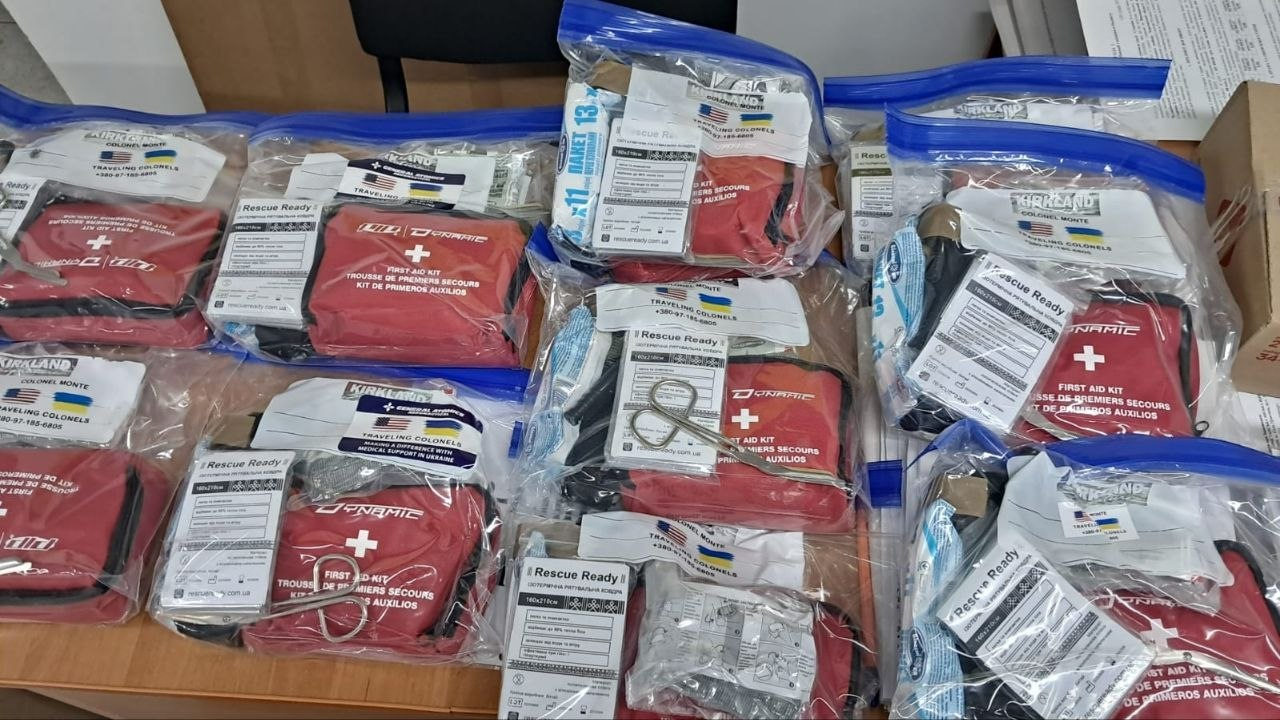 Великий набір індивідуальних тактичних аптечок першої допомоги, розкладених на столі. Кожен медичний набір складається з червоного підсумка з білим хрестом і написів англійською, французькою та іспанською мовами, а також додаткових засобів. Комплекти упаковані у прозорі пластикові пакети із зіп-замком, на яких наклеєні стікери з прапорами США та України й назвою «COLONEL MONTE».