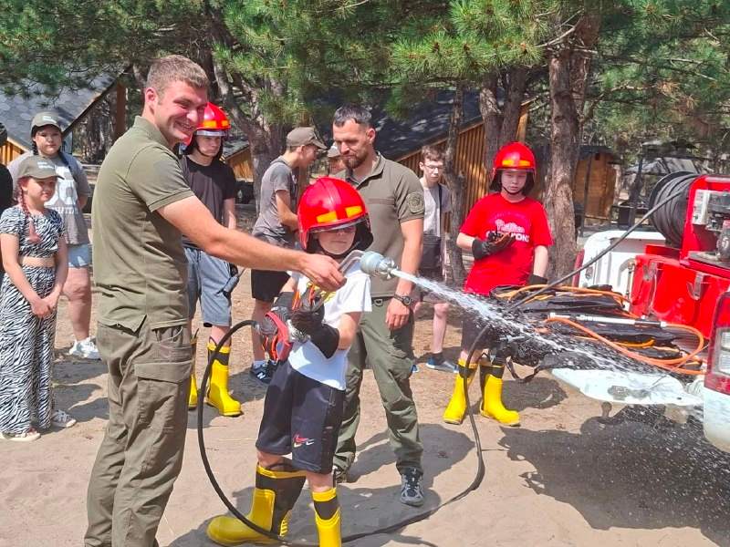 Чоловік у військовій або службовій формі хакі допомагає хлопчикові в червоній пожежній касці та жовтих гумових чоботях тримати пожежний рукав і пускати воду з кузова рятувального автомобіля. За ними спостерігають інші діти та інструктори на лісовому майданчику на тлі дерев і дерев'яних будиночків.