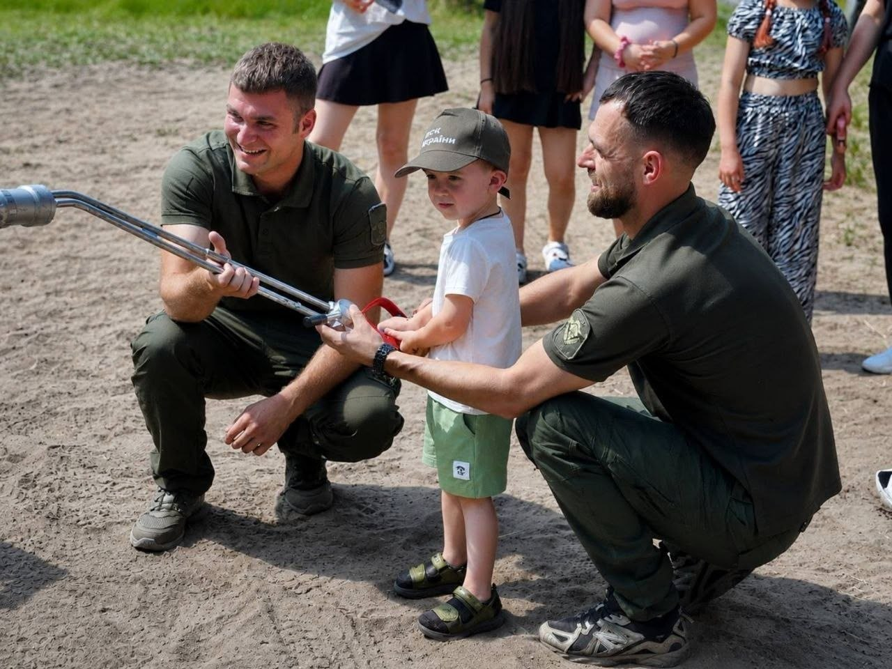 Двоє чоловіків у військовій або службовій формі кольору хакі присіли поруч із маленьким хлопчиком у зеленій кепці та допомагають йому тримати металевий пожежний ствол. Усі троє усміхаються, а на задньому плані на піщаному майданчику стоять інші діти та дорослі.