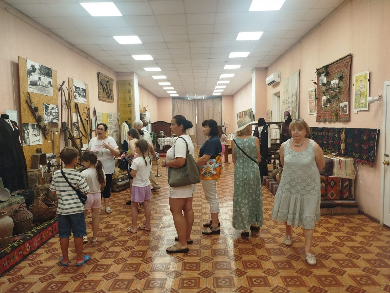 Група людей, серед яких є дорослі та діти, оглядає експонати в довгій музейній залі з візерунчастою підлогою. На стінах та спеціальних стендах розміщені предмети побуту, старовинні знаряддя праці, світлини, ткані килими та традиційний одяг.