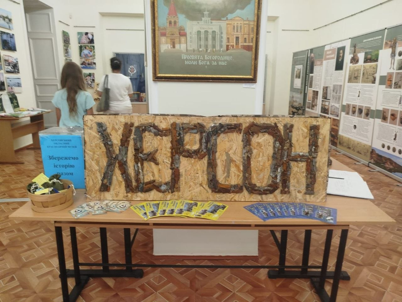 Великий дерев'яний щит із написом «ХЕРСОН», викладеним з уламків чи кори, стоїть на столі в музейній залі. Поруч розкладені інформаційні буклети та синя благодійна скринька з написом «Збережемо історію разом», а на задньому плані видно музейні стенди, картину та відвідувачів.