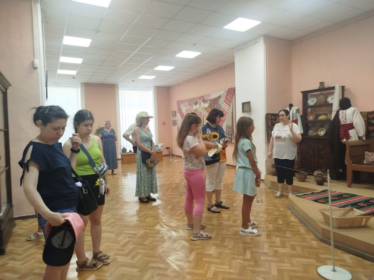 Група відвідувачів, серед яких дорослі та діти, слухає екскурсовода у світлій музейній залі. Праворуч на підвищенні облаштовано етнографічну експозицію з традиційним українським одягом, автентичними меблями, посудом та тканим килимом.