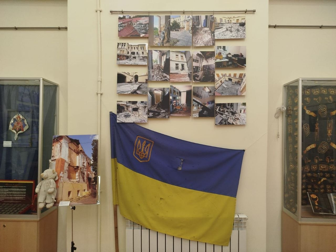 Музейна експозиція, присвячена руйнуванням внаслідок війни. На світлій стіні розвішано колаж із фотографій зруйнованих будівель та кабінетів, під якими закріплено великий державний прапор України з тризубом та слідами пошкоджень. По обидва боки розташовані скляні вітрини з експонатами, а ліворуч на стійці стоїть знімок понівеченого будинку та м'яка іграшка.