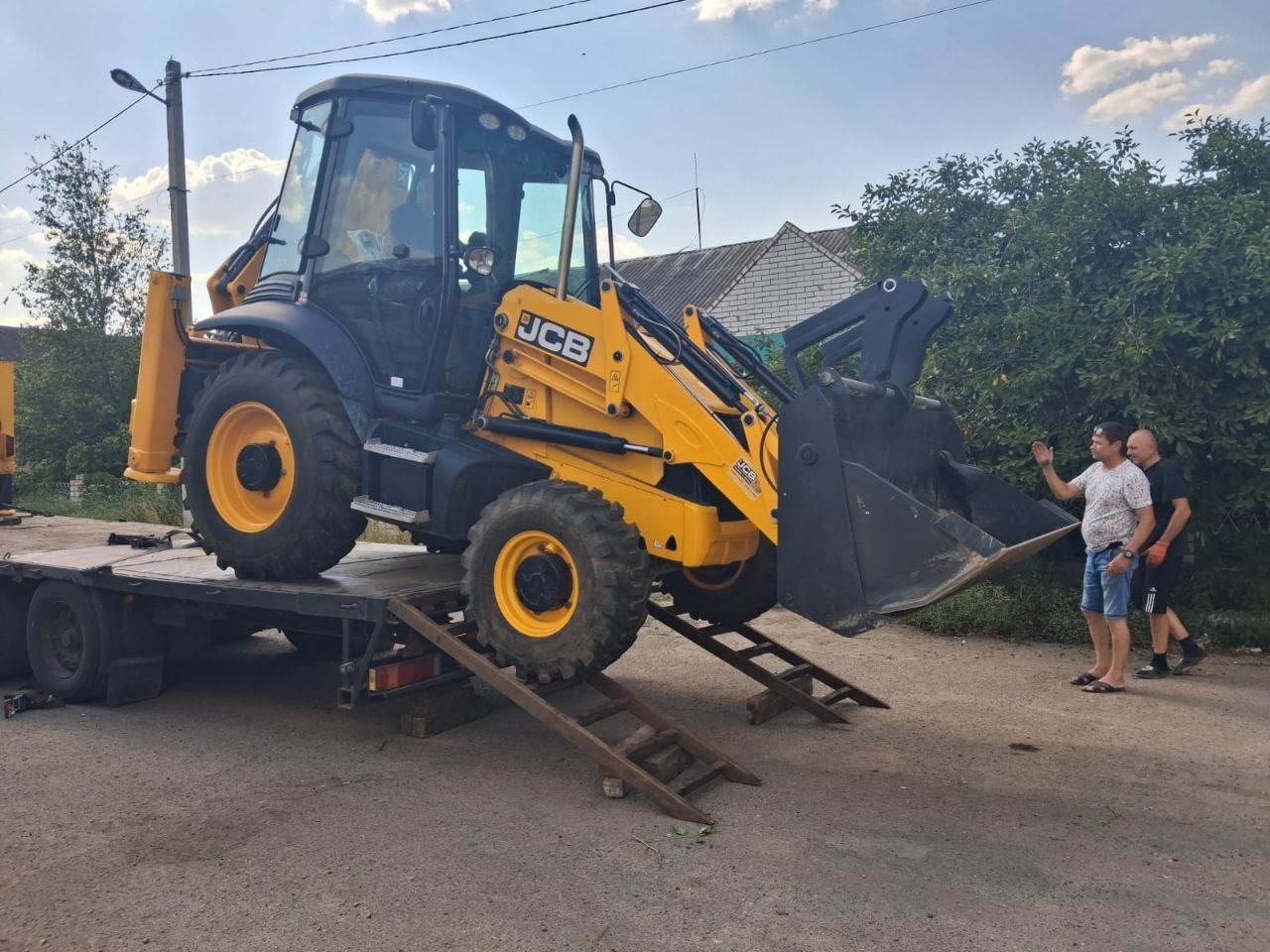 Жовтий екскаватор-навантажувач марки JCB заїжджає по металевих апарелях на платформу вантажного причепа. Праворуч на вулиці стоять двоє чоловіків у повсякденному одязі, один з яких жестикулює рукою, допомагаючи координувати процес завантаження спецтехніки на тлі зелених дерев і житлових будинків.