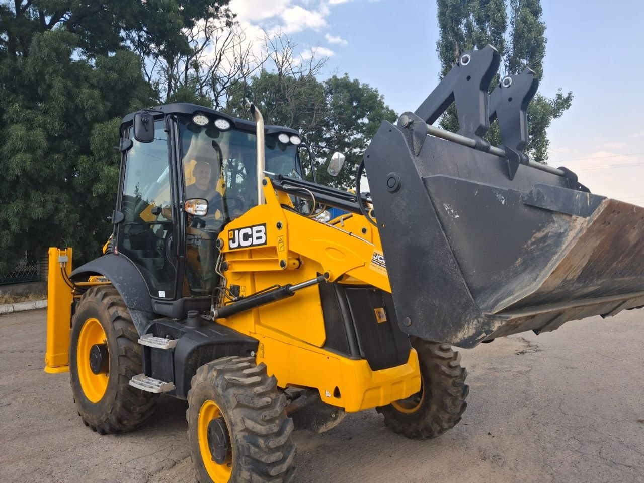 Жовтий екскаватор-навантажувач марки JCB стоїть на асфальтованому майданчику з піднятим великим переднім ковшем. У скляній кабіні спецтехніки видно силует водія, а на задньому плані височіють зелені дерева під світлим небом із легкими хмарами.