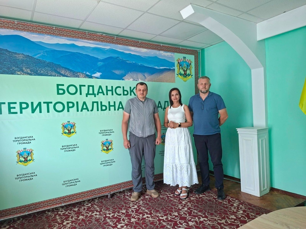 Двоє чоловіків та одна жінка стоять у приміщенні на тлі великого світло-зеленого банера з написом «БОГДАНСЬКА ТЕРИТОРІАЛЬНА ГРОМАДА», зображенням гір та гербами. Чоловік ліворуч одягнений у сіру сорочку та штани, жінка по центру — у білу довгу сукню, а чоловік праворуч — у синю футболку-поло та темні штани.