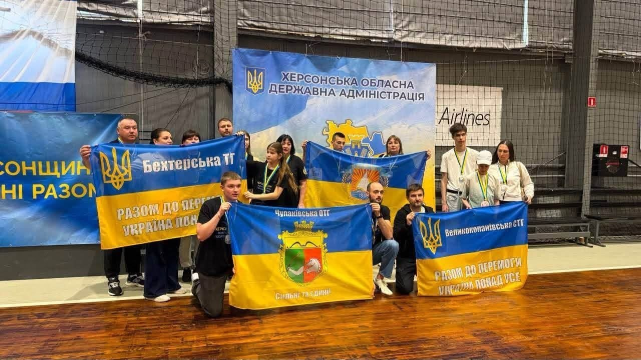 Група людей стоїть і сидить на дерев'яному спортивному паркеті у спортивній залі, тримаючи три великі синьо-жовті прапори з гербами та написами «Бехтерська ТГ», «Чулаківська ОТГ» та «Великокопанівська СТГ». На задньому плані розміщено великий банер «Херсонська обласна державна адміністрація», а учасники заходу мають на шиях медалі.