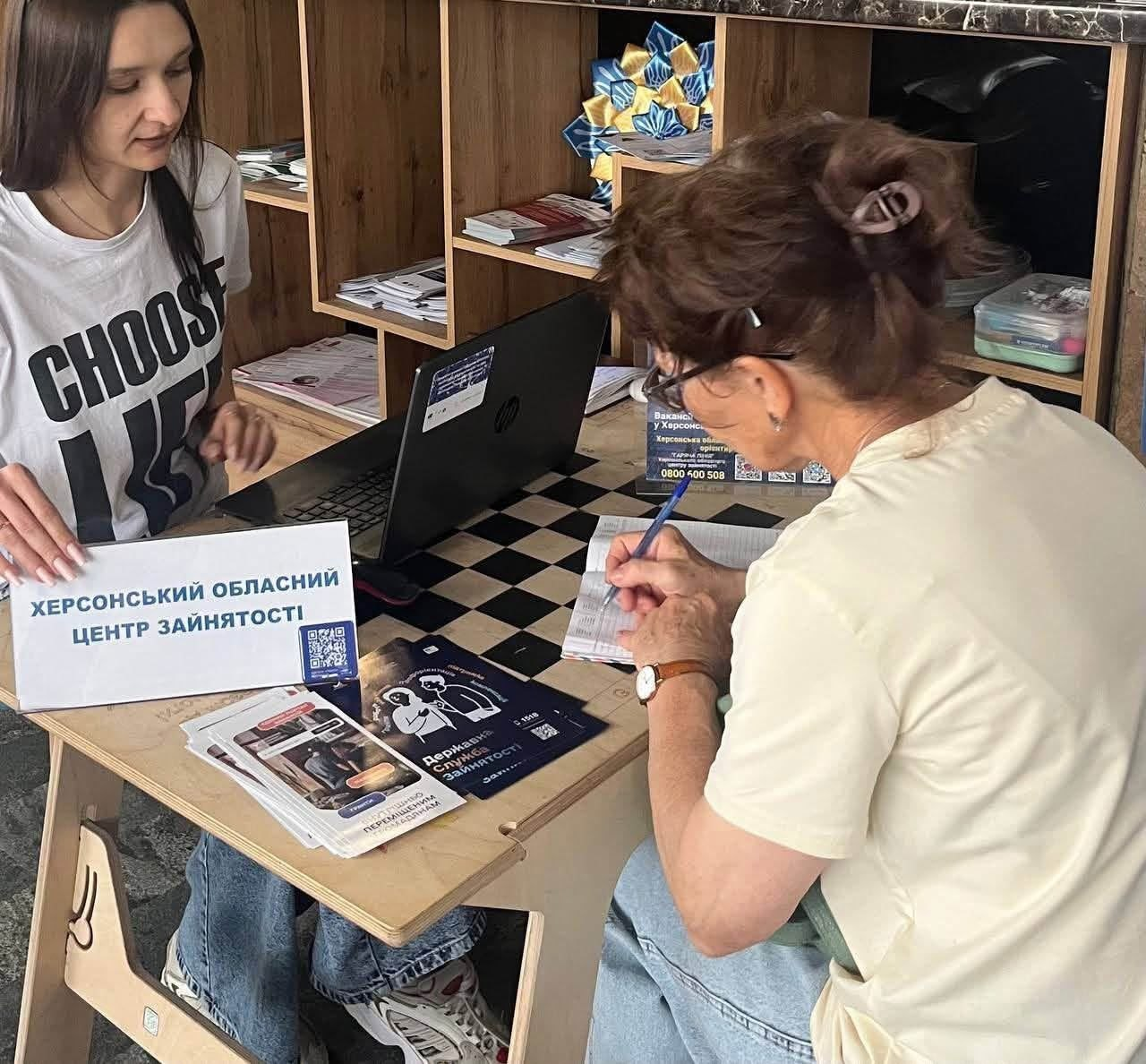 Дві жінки сидять за невеликим столиком у приміщенні під час консультації. Жінка ліворуч у білій футболці з написом дивиться на документи, поряд із якими стоїть табличка «ХЕРСОНСЬКИЙ ОБЛАСНИЙ ЦЕНТР ЗАЙНЯТОСТІ». Жінка праворуч, повернута спиною до камери, робить записи ручкою в блокноті на тлі ноутбука, брошур та дерев'яних полиць.