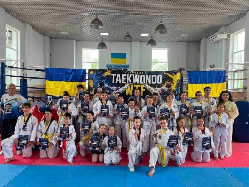 Велика група дітей та підлітків у білих костюмах для тхеквондо (добок) з різноколірними поясами та медалями стоїть на спортивному маті (татамі) у залі. Майже кожен тримає в руках нагородну грамоту. На задньому плані по обидва боки висять державні прапори України, а в центрі розміщено великий темний банер із написом «TAEKWONDO WTF».