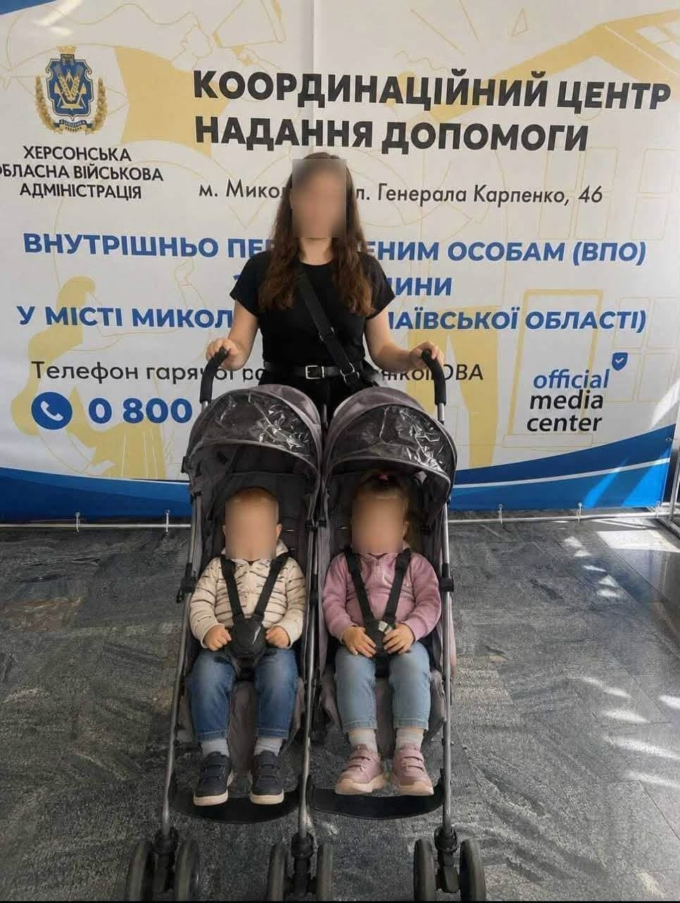 Жінка в чорній футболці стоїть позаду подвійної дитячої коляски, у якій сидять двоє маленьких дітей — хлопчик у світлій кофті та дівчинка в рожевій куртці. Обличчя жінки та дітей штучно приховані розмиттям задля конфіденційності. Вони перебувають на тлі великого інформаційного банера з написом «КООРДИНАЦІЙНИЙ ЦЕНТР НАДАННЯ ДОПОМОГИ» та символікою Херсонської ОВА.