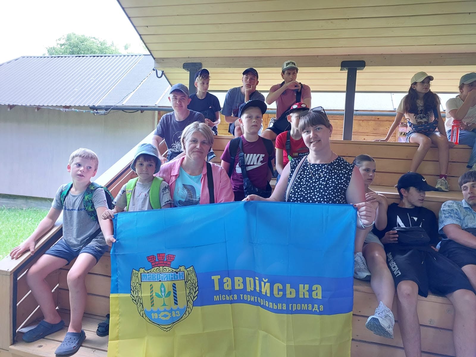 Група дітей та дорослих сидить на дерев'яних сходах і трибунах просто неба. Дві жінки на передньому плані тримають великий синьо-жовтий прапор із гербом та написом «Таврійська міська територіальна громада».