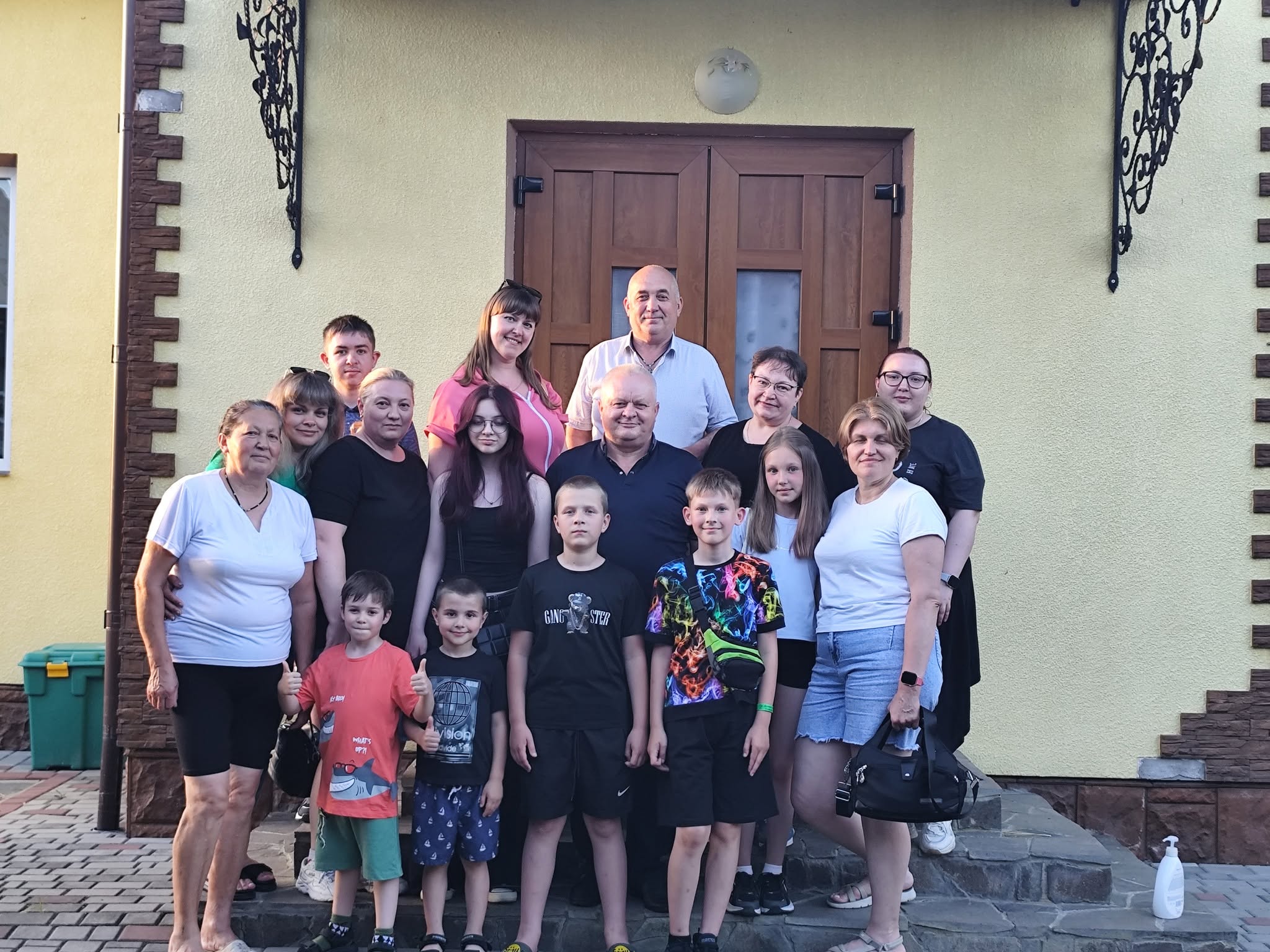 Велика група дорослих та дітей у літньому повсякденному одязі позирує для спільного фото на кам'яному ганку перед дерев'яними дверима будівлі.